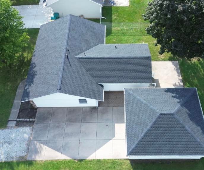 An aerial drone view of a completed residential roofing project in a suburban neighborhood featuring dark gray architectural shingles and clean roof lines.