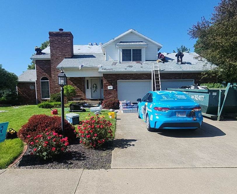 Rainforcing Roofing team members performing a roof replacement on a large brick and siding home, with a branded blue company vehicle and a large debris dumpster on-site.