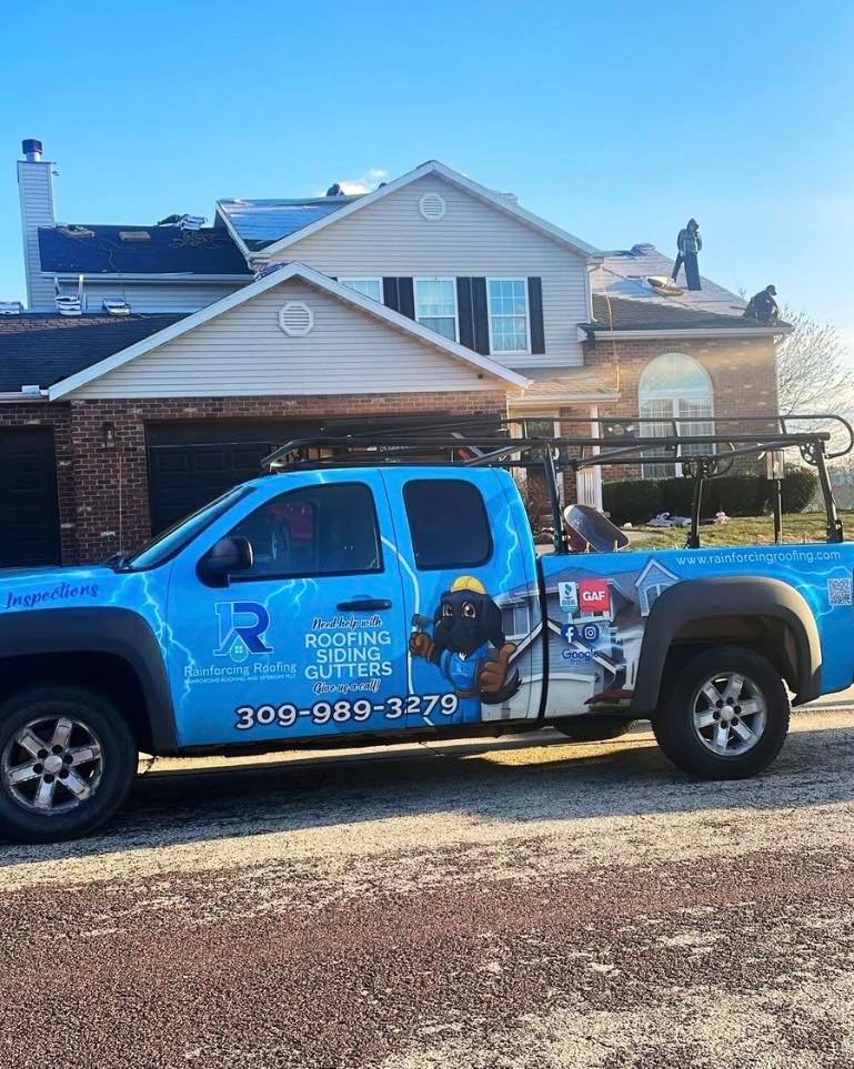 A branded company service truck with a ladder rack and "Rainforcing Roofing" graphics parked at a residential job site. The truck prominently displays the company phone number (309) 989-3279 and GAF certification logo while a crew works on the roof in the background.