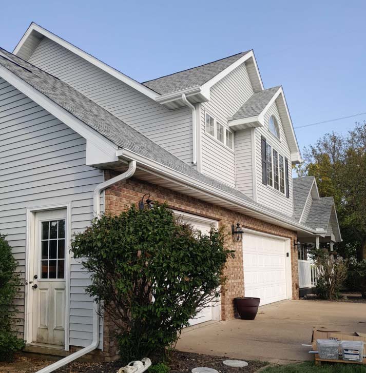 A side view of a large residential home featuring newly installed light gray horizontal vinyl siding, crisp white soffit and fascia, and a matching white gutter and downspout system. The lower portion of the home combines this fresh siding with traditional red brick accents near the garage and side entrance.